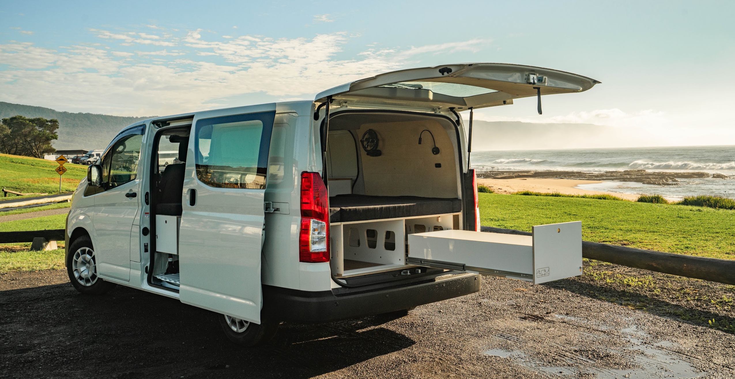 Toyota Hiace amper van with open rear door parked by a scenic coastal area.